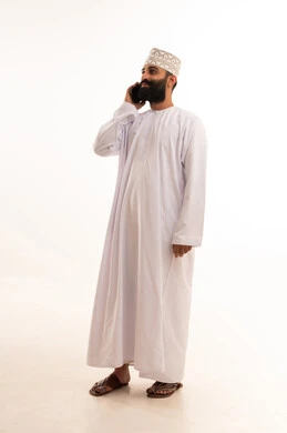 A portrait of an Arab Gulf Omani man wearing a dishdasha and an Omani kumma, wearing prescription glasses, holding a mobile phone in his hand and making a phone call, using modern and advanced technologies, gestures of happiness upon hearing good news, full-body shot, white background.