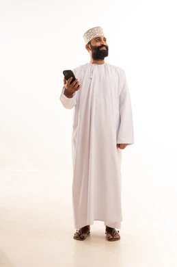 Gestures of happiness upon hearing good news, using modern and advanced technologies, a portrait of an elderly Arab Gulf Omani man wearing a dishdasha and an Omani kumma, wearing reading glasses, holding a mobile phone in his hand and making a phone call, full-body shot, white background.