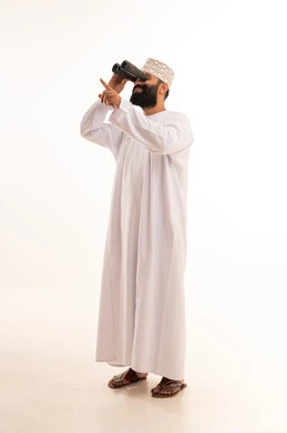 The love of adventure and exploration, seeing distant things through binoculars, a portrait of a young Arab Gulf Omani man wearing a dishdasha and kumma looking through a binocular, pointing with his index finger at something with gestures of joy and happiness, white background.