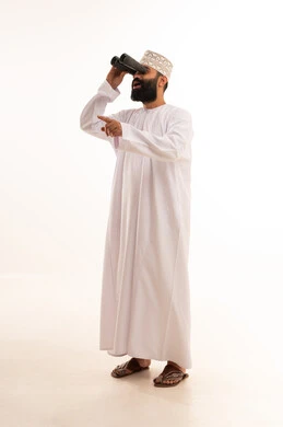 رؤية الاشياء البعيدة عبر المنظار، حب المغامرة والاستكشاف، بورتريه لشاب عربي خليجي عماني يرتدي الدشداشة والكمه ينظر من خلال المنظور ثنائي العينين يشير بإصبع السبابة الى شيء ما بإيماءات الفرح والسرور، خلفية بيضاء