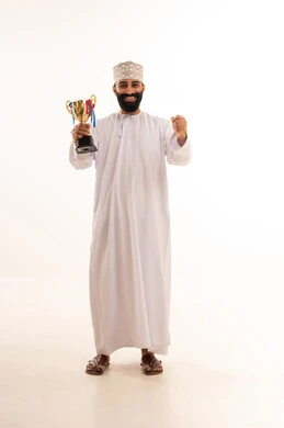 A portrait of an Arab Gulf Omani man wearing a dishdasha and kumma sitting on a chair holding a golden cup, looking at the camera with gestures of joy and enthusiasm, celebrating the championship and victory, achieving first place in the competition, with a white background.