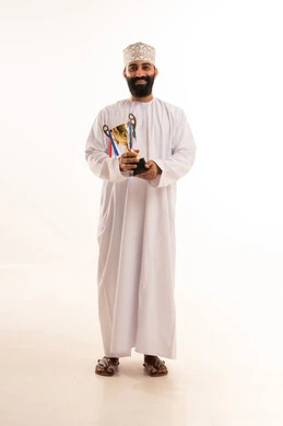الحصول على المركز الأول في المسابقة، التتويج بالبطولة والفوز، بورتريه لرجل عربي خليجي عماني يرتدي الدشداشه والكمه يجلس على كرسي يحمل بيده الكأس الذهبية ينظر إلى الكاميرا بايماءات السرور والحماس، خلفية بيضاء