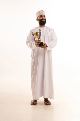 التتويج بالبطولة والفوز، الحصول على المركز الأول في المسابقة، بورتريه لرجل عربي خليجي عماني يرتدي الدشداشه والكمه يجلس على كرسي يحمل بيده الكأس الذهبية ينظر إلى الكاميرا بايماءات السرور والحماس، خلفية بيضاء