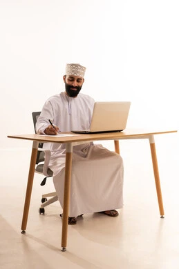 A youthful Omani job and profession, using a modern advanced technical device, a portrait of an Arab Gulf Omani man wearing a kumma and dishdasha sitting on a chair in front of a wooden table writing down ideas and recording information on white papers, white background.
