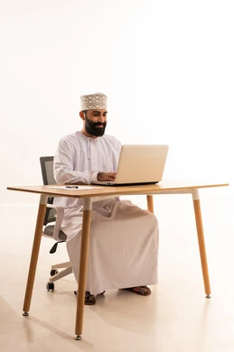 The use of advanced technology, focusing on task completion, integrating modern technologies in the administrative and office field, working in Omani offices and companies, a portrait of an Arab Gulf Omani man wearing a dishdasha and kumma sitting on an office chair and working on a laptop, white background.