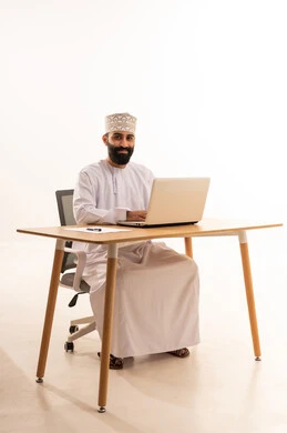 Focusing on task completion, integrating modern technologies in the administrative and office field, working in Omani offices and companies, a portrait of an Arab Gulf Omani man wearing a dishdasha and kumma sitting on an office chair and working on a laptop, white background.