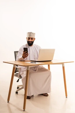 العمل في المكاتب والشركات العمانية، تصفح مواقع التواصل الاجتماعي ، اجراء مكالمة هاتفية عبر الجوال ، التركيز في إنجاز المهام، دمج التقنيات الحديثة بالمجال الإداري والمكتبي،بورتريه لرجل عربي خليجي عماني يرتدي الدشداشه والكمة يجلس على كرسي المكتب ويعمل على جهاز الحاسوب المحمول، خلفية بيضاء