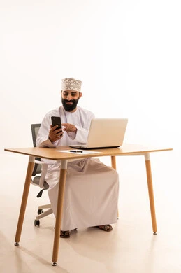 التركيز في إنجاز المهام، تصفح مواقع التواصل الاجتماعي ، اجراء مكالمة هاتفية عبر الجوال ، دمج التقنيات الحديثة بالمجال الإداري والمكتبي، العمل في المكاتب والشركات العمانية، بورتريه لرجل عربي خليجي عماني يرتدي الدشداشه والكمة يجلس على كرسي المكتب ويعمل على جهاز الحاسوب المحمول، خلفية بيضاء