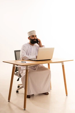 Browsing social media, making a phone call via mobile, focusing on completing tasks, integrating modern technologies in the administrative and office field, working in Omani offices and companies, a portrait of an Arab Gulf Omani man wearing a dishdasha and kumma sitting on an office chair and working on a laptop, white background.