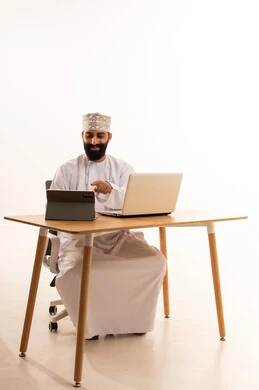 Conducting a live broadcast via the tablet, watching movies and series with expressions of happiness, integrating modern technologies in work, a portrait of an Arab Gulf Omani man wearing a dishdasha and a kumma sitting on an office chair working on a laptop, white background.