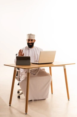 دمج التقنيات الحديثة فى العمل، اجراء بث مباشر عبر الجهاز اللوحى  ، مشاهده الافلام والمسلسلات بتعابير السعاده ،بورتريه لرجل عربي خليجي عماني يرتدي الدشداشه والكمة يجلس على كرسي المكتب يعمل على جهاز الحاسوب المحمول ، خلفية بيضاء