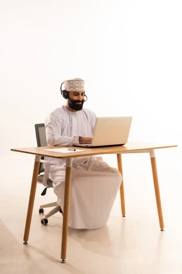 بورتريه لرجل عربي خليجي عماني يرتدي الدشداشه والكمة والهيدفون يجلس على كرسي المكتب ويعمل على جهاز الحاسوب المحمول، التركيز في إنجاز المهام، دمج التقنيات الحديثة بالمجال الإداري والمكتبي، سماع الموسيقي عبر سماعات الرأس، العمل في المكاتب والشركات العمانية، خلفية بيضاء