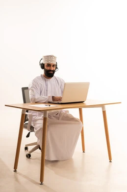 Focusing on task completion, integrating modern technologies in the administrative and office field, listening to music through headphones, working in Omani offices and companies, a portrait of an Arab Gulf Omani man wearing a dishdasha and kumma with headphones sitting on an office chair working on a laptop, white background.