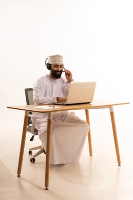 العمل في المكاتب والشركات العمانية، بورتريه لرجل عربي خليجي عماني يرتدي الدشداشه والكمة والهيدفون يجلس على كرسي المكتب يعمل على جهاز الحاسوب المحمول يقوم بتدوين الأفكار وتسجيل المعلومات على الأوراق البيضاء، التركيز في إنجاز المهام، دمج التقنيات الحديثة بالمجال الإداري والمكتبي، سماع الموسيقي عبر سماعات الرأس، خلفية بيضاء