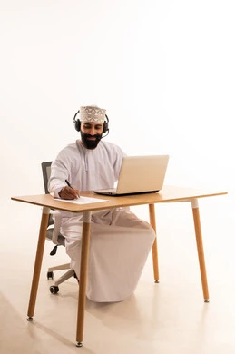 Listening to music through headphones, focusing on completing tasks, integrating modern technologies in the administrative and office field, working in Omani offices and companies, a portrait of an Arab Gulf Omani man wearing a dishdasha and kumma with headphones sitting on an office chair working on a laptop, jotting down ideas and recording information on white papers, white background.
