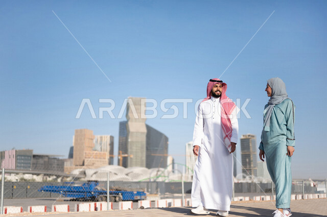 زوجان عربيان خليجيان سعوديان سعيدان يمارسان رياضة المشي في إحدي منتزهات الرياض ، الاستجمام و التنزه في الهواء الطلق ، كامل الجسم ، صورة بالطول الكامل ، قضاء وقت ممتع في مدينة الملك عبد الله المالية ( كافد ) في مدينة الرياض ، المملكة العربية السعودية