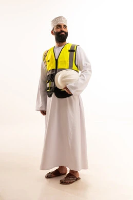 العمل في القطاع الهندسي، مفهوم الهندسة والانشاءات المعمارية، بورتريه لمهندس عربي خليجي عماني يرتدي الدشداشة والكمة وسترة الحماية يمسك بيده الخوذة البيضاء بتعابير السرور، مهنة ووظيفة للشباب في سلطنة عمان، الجسم بالطول الكامل، خلفية بيضاء