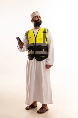 A portrait of an Arab Gulf Omani engineer wearing a dishdasha, a kumma, and a safety vest, holding a mobile phone in his hand, browsing social media, using modern technologies in the engineering sector, integrating advanced technology into work, full-body shot, white background.