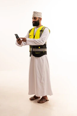 Integrating advanced technology into work, browsing social media, using modern technologies in the engineering sector, a portrait of an Arab Gulf Omani engineer wearing a dishdasha and kumma and a safety vest holding a mobile phone in his hand, full-body, white background.
