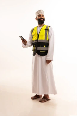 استخدام التقنيات الحديثة بالقطاع الهندسي، تصفح مواقع التواصل الاجتماعي، دمج التكنولوجيا المتطورة بالعمل، بورتريه لمهندس عربي خليجي عماني يرتدي الدشداشة والكمة وسترة الحماية يمسك الجوال بيده، الجسم بالطول الكامل، خلفية بيضاء