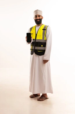 تصفح مواقع التواصل الاجتماعي، استخدام التقنيات الحديثة بالقطاع الهندسي، دمج التكنولوجيا المتطورة بالعمل، بورتريه لمهندس عربي خليجي عماني يرتدي الدشداشة والكمة وسترة الحماية يمسك الجوال بيده، الجسم بالطول الكامل، خلفية بيضاء
