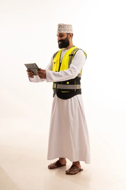 مفهوم الهندسة والانشاءات المعماريه، إدارة المشاريع بحرفية باستخدام الجهاز اللوحي ،دمج التقنيات الحديثة ، العمل في القطاع الهندسي، بورتريه  لمهندس عربي خليجي عماني يرتدي الدشداشة والكمة وسترة الحماية يمسك بيده التابلت ، خلفية بيضاء
