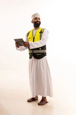 العمل في القطاع الهندسي، إدارة المشاريع بحرفية باستخدام الجهاز اللوحي ،دمج التقنيات الحديثة ، مفهوم الهندسة والانشاءات المعماريه، بورتريه  لمهندس عربي خليجي عماني يرتدي الدشداشة والكمة وسترة الحماية يمسك بيده التابلت ، خلفية بيضاء
