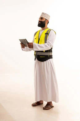 دمج التقنيات الحديثة ، إدارة المشاريع بحرفية باستخدام الجهاز اللوحي ،العمل في القطاع الهندسي، مفهوم الهندسة والانشاءات المعماريه، بورتريه  لمهندس عربي خليجي عماني يرتدي الدشداشة والكمة وسترة الحماية يمسك بيده التابلت ، خلفية بيضاء