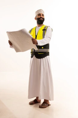 مفهوم الانشاءات المعمارية، بورتريه لمهندس عربي خليجي عماني يرتدي الدشداشة والكمة وسترة الحماية يمسك بيديه المخطط، العمل في مجال الهندسة، مهنة ووظيفة للشباب في سلطنة عمان، الإشراف على مخططات المشاريع الهندسية، الجسم بالطول الكامل، خلفية بيضاء