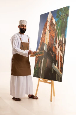 مفهوم الفن والالوان ،استخدام ريشة التلوين، أنشطة ترفيهية فنية ممتعة، بورتريه لرسام عربي خليجي عماني يرتدي الدشداشة والكمة وقبعة يحمل بيديه الالوان المائية, ورشة عمل وفنون, خلفية بيضاء