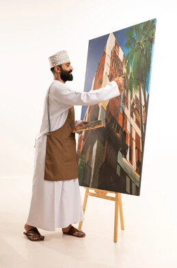 ورشة عمل وفنون, استخدام ريشة التلوين، أنشطة ترفيهية فنية ممتعة، بورتريه لرسام عربي خليجي عماني يرتدي الدشداشة والكمة وقبعة يحمل بيديه الالوان المائية,خلفية بيضاء