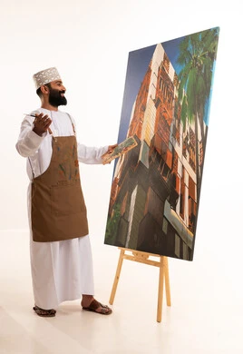 بورتريه لرسام عربي خليجي عماني يرتدي الدشداشة والكمة وقبعة يحمل بيديه الالوان المائية, ورشة عمل وفنون, استخدام ريشة التلوين، أنشطة ترفيهية فنية ممتعة، خلفية بيضاء