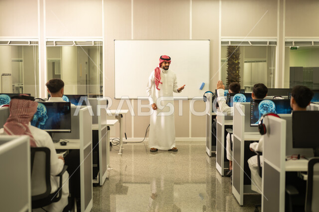 مفهوم الاجتهاد وطلب التفوق، معلم عربي خليجي سعودي يرتدي الثوب التقليدي والشماغ يقوم بمناقشة الدروس مع طلابه, التعليم فى المملكة العربية السعودية, طلاب عرب يجلسون أمام أجهزة الكومبيوتر في مختبر الحاسوب
