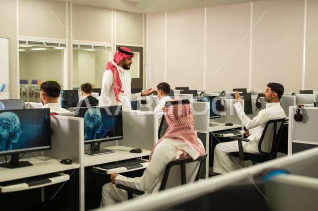 Digital learning in schools: A group of Saudi Arabian Gulf Arab students sit in front of a computer listening to a lecture. A Saudi Arabian Gulf Arab university professor, wearing a thobe and shemagh, explains to his students in the computer lab.