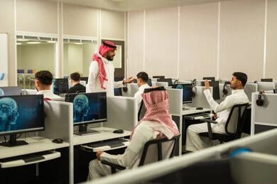 Digital learning in schools: A group of Saudi Arabian Gulf Arab students sit in front of a computer listening to a lecture. A Saudi Arabian Gulf Arab university professor, wearing a thobe and shemagh, explains to his students in the computer lab.