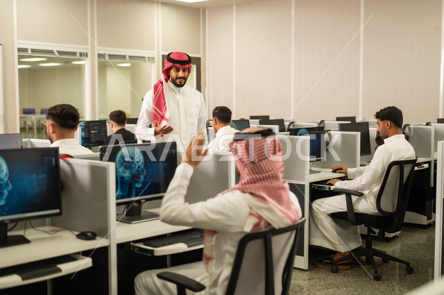 Student participation by raising hands; a Saudi Arabian teacher wearing traditional thobe and shemagh explains and provides training on computer equipment; studying and learning in the Kingdom of Saudi Arabia.