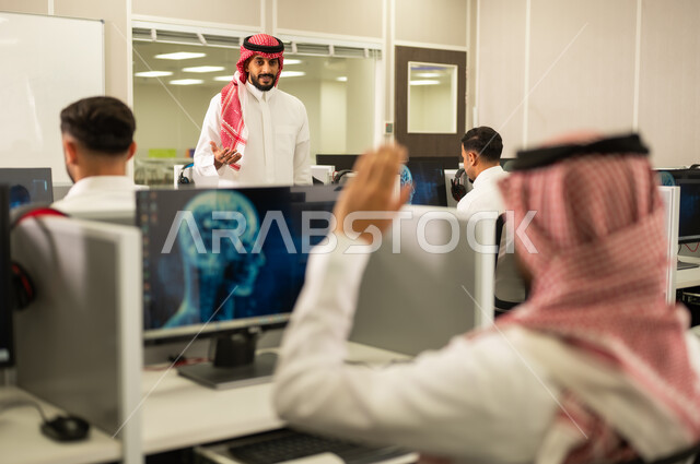 الاجتهاد الأكاديمي لتحقيق التميز, طالب عربى خليحى سعودى يتفاعل في المحاضرة من خلال رفع يديه للاجابة على اسئلة المدرس, معلم  يرتدي الثوب التقليدي والشماغ يقوم بمناقشة الدروس مع طلابة فى مختبر الحاسوب