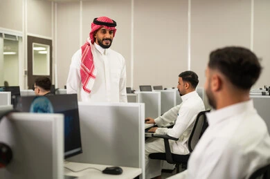 Using computers for school learning: A group of Saudi Arabian students wearing traditional attire sit in front of computers in a modern lab. Education in Saudi Arabia: A group of employees work in front of computers.