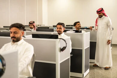 Colleague participation and collaboration; education in Saudi Arabia; Arab students sitting in front of computers in a modern lab; a group of Saudi Arabian Gulf employees wearing traditional attire working at computers in the company headquarters.