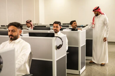 Advanced colleges and universities; a group of Arab Gulf and Saudi students wearing traditional attire sit in front of computers in a modern lab, interacting and participating within the classroom.