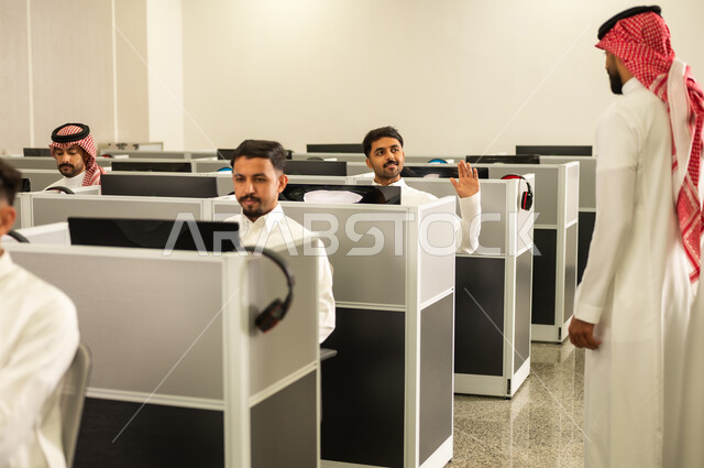 الدراسة الرقمية في المدارس، مجموعة من  الطلاب العرب الخليجيون السعوديون يجلسون امام الحاسوب يقومون بالإستماع الى المحاضره، أستاذ جامعي عربي خليجي سعودى يرتدي الثوب و الشماغ يشرح لطلابه في الفصل الدراسى