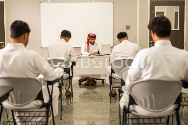 A Saudi Arabian teacher, wearing traditional thobe and shemagh, explains a lesson to students using a computer, demonstrating clear and concise explanations and effective communication. The lesson is interactive, engaging, and participatory within the classroom.