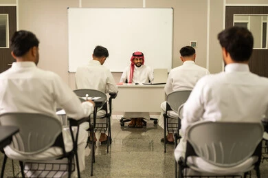 Enhancing scientific knowledge and fostering creativity; observing a group of students in a classroom; an Arab Gulf Saudi teacher wearing traditional thobe and shemagh explaining concepts to his students in a modern school environment; the modern education system; quality of learning in academic schools.