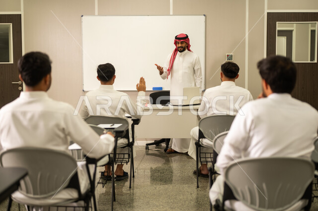 التربية والتعليم بالمناهج المقررة للطلاب في المملكة العربية السعودية, طلاب عرب خليجيون سعوديون يستمعون الى الدرس, معلم عربي خليجي سعودي يرتدي الثوب التقليدي والشماغ يقوم بمناقشة الدروس مع طلابه باستخدام الحاسوب