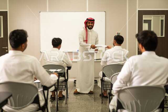 Striving for excellence, a group of Saudi Arabian Gulf students prepare to answer exam questions. Education in the Kingdom of Saudi Arabia: A Saudi Arabian Gulf teacher, wearing a traditional thobe and shemagh, distributes exam papers to the students.