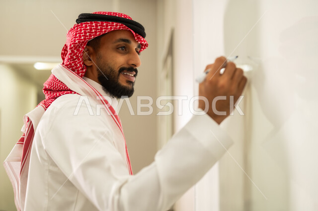 استخدام السبورة لسهولة الشرح, المقررات والمناهج التعليمية الحديثة والمطورة بالمملكة العربية السعودية, الاهتمام بقدرات الطلاب بتقديم الدروس, معلم عربي خليجي سعودي يرتدي الثوب التقليدي والشماغ يقوم بالشرح على اللوح الأبيض