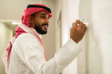 Using the whiteboard for ease of explanation; modern and developed educational curricula and programs in the Kingdom of Saudi Arabia; attention to students' abilities in lesson delivery; a Saudi Arabian Gulf Arab teacher wearing the traditional thobe and shemagh explaining on the whiteboard.