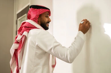 المقررات والمناهج التعليمية الحديثة والمطورة بالمملكة العربية السعودية, الاهتمام بقدرات الطلاب بتقديم الدروس, معلم عربي خليجي سعودي يرتدي الثوب التقليدي والشماغ يقوم بالشرح على اللوح الأبيض، استخدام السبورة لسهولة الشرح