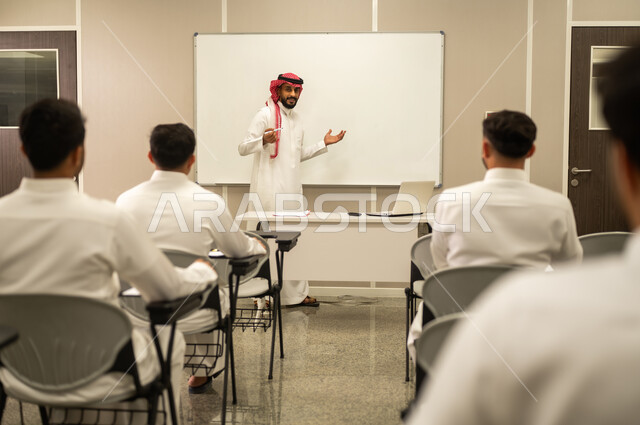 The importance of science in developing mental abilities; educational curricula and syllabi in the Kingdom of Saudi Arabia; a photograph from behind of Saudi Arabian Gulf students sitting at their school desks listening to their teacher during a lesson; gestures of focus and concentration; use of the whiteboard.