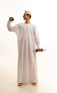 Lifting heavy weights is a physical activity to maintain physical fitness, build and strengthen body muscles. A portrait of an Arab Gulf Omani man wearing a dishdasha and kumma, with glasses, holding weights in his hand, against a white background.